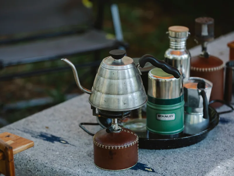 Gooseneck kettle on a camp stove with coffee gear on a picnic table