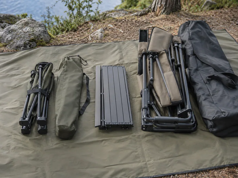 Folded camping chair, compact side table and carrying bags laid beside a larger packed chair